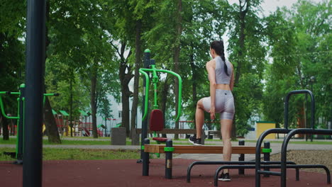 A-young-woman-in-a-Park-performs-striding-lunges-on-a-bench-in-sportswear-in-the-summer.-Athletics-Caucasian-woman-trains-in-the-Park.
